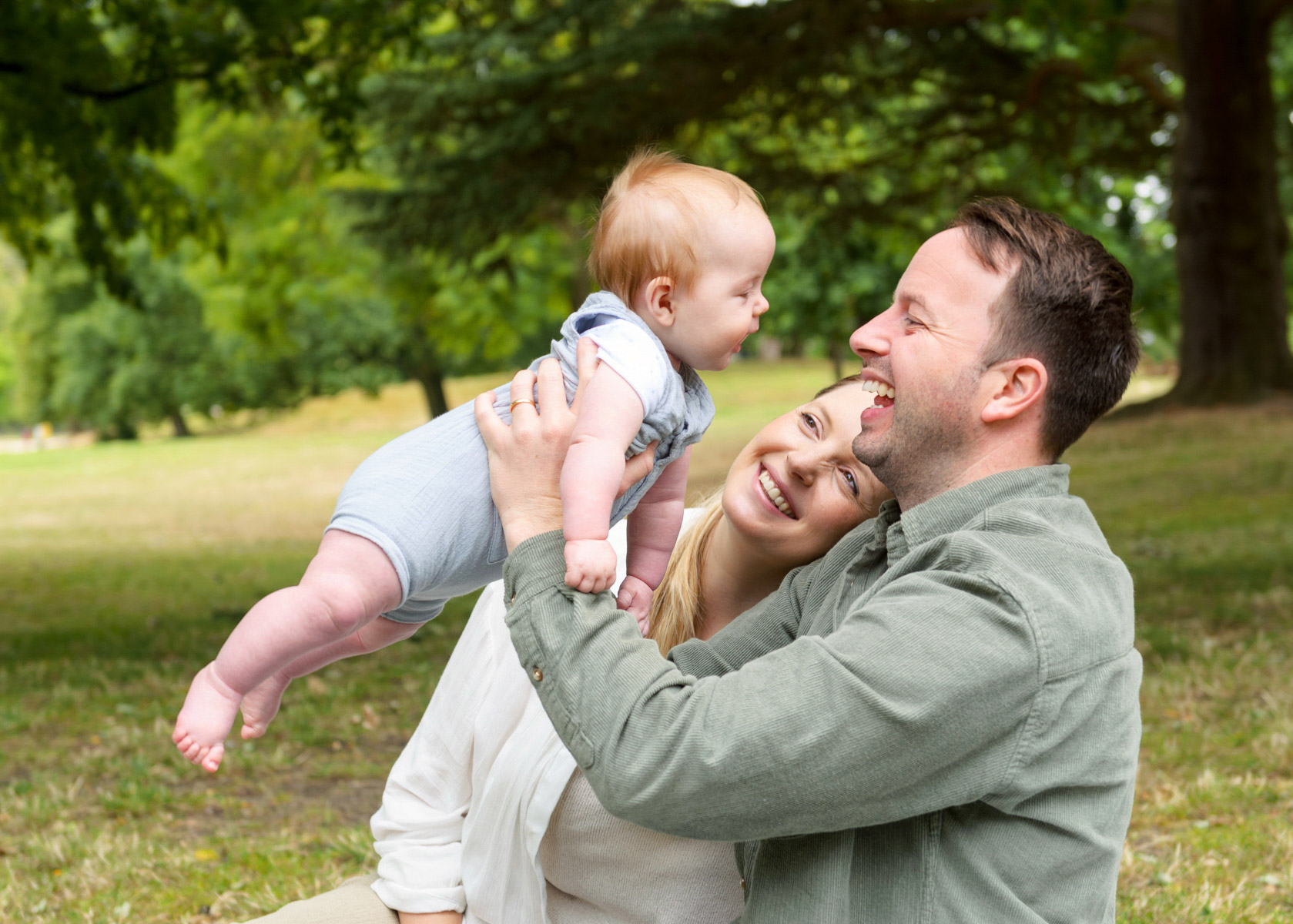 family photo of a dad holding up newborn baby with mum sitting on grass near the dinosaurs in Crystal Palace park South London