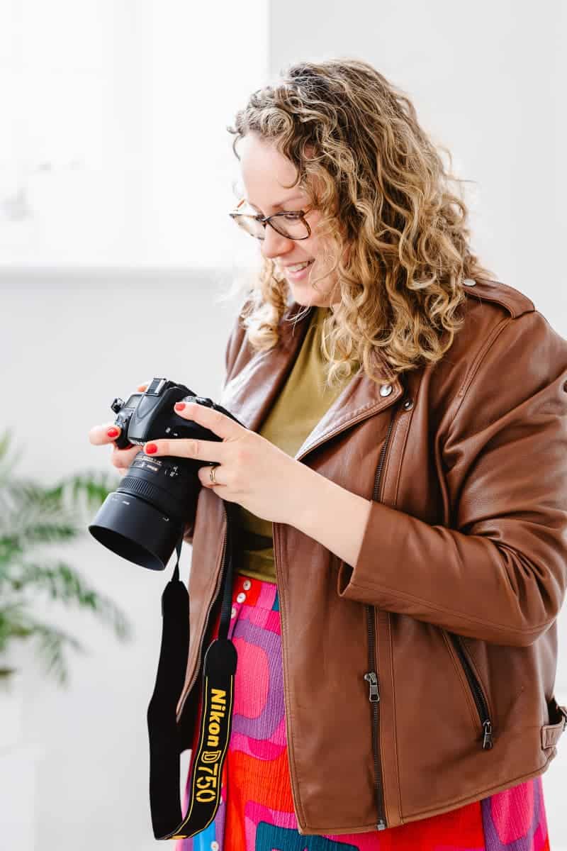 A portrait of Molly Rhianon looking at the back of her camera during a photoshoot. Molly wears a brown leather jacket over a bright red patterned skirt and has curly hair. 