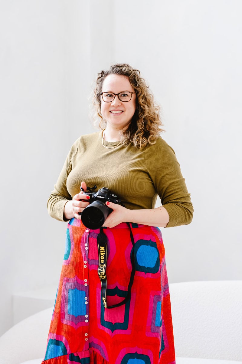 a portrait photograph of Molly Rhianon Behag who is the founder of Molly Rhianon Photography. Molly stands confidently against a white background holding a camera and wearing olive green top and bright red and pink patterned skirt.