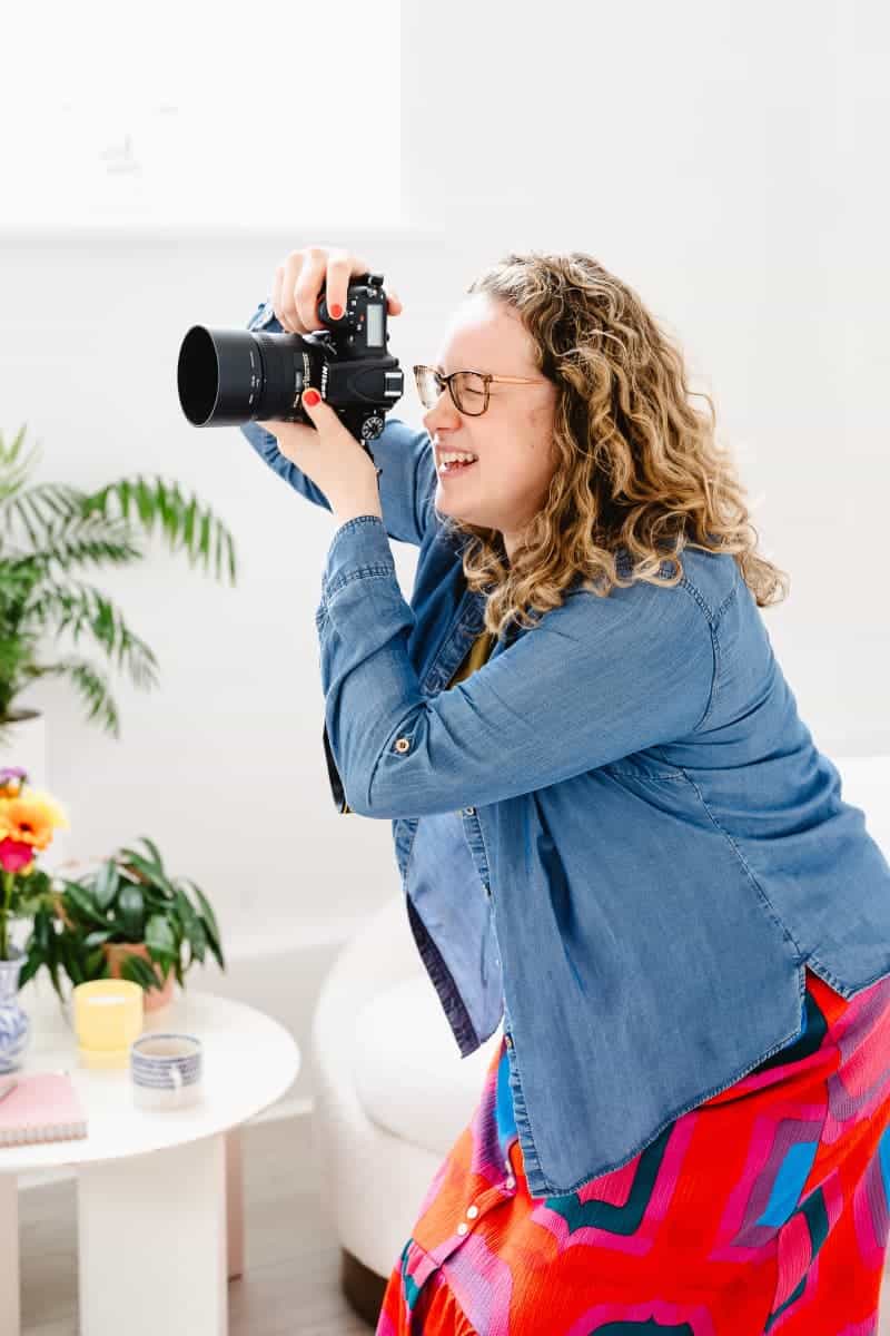Molly Rhianon squatting while photographing and holding the camera up to her face. She wears a denim shirt and bright red patterned skirt and has plants and flowers visible behind her.