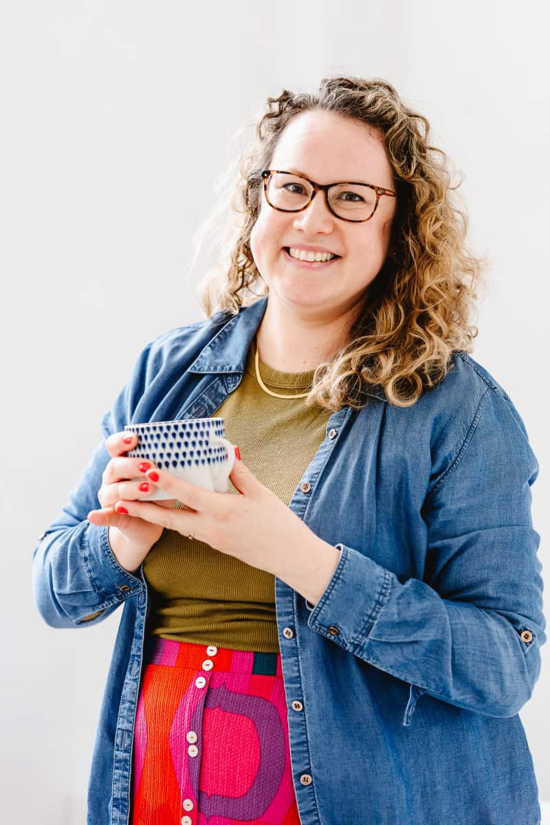 a portrait photograph of Molly Rhianon Behag who is the founder of Molly Rhianon Photography. Molly stands smiling against a white background holding a blue and white mug of coffee and wearing olive green top and bright red and pink patterned skirt.