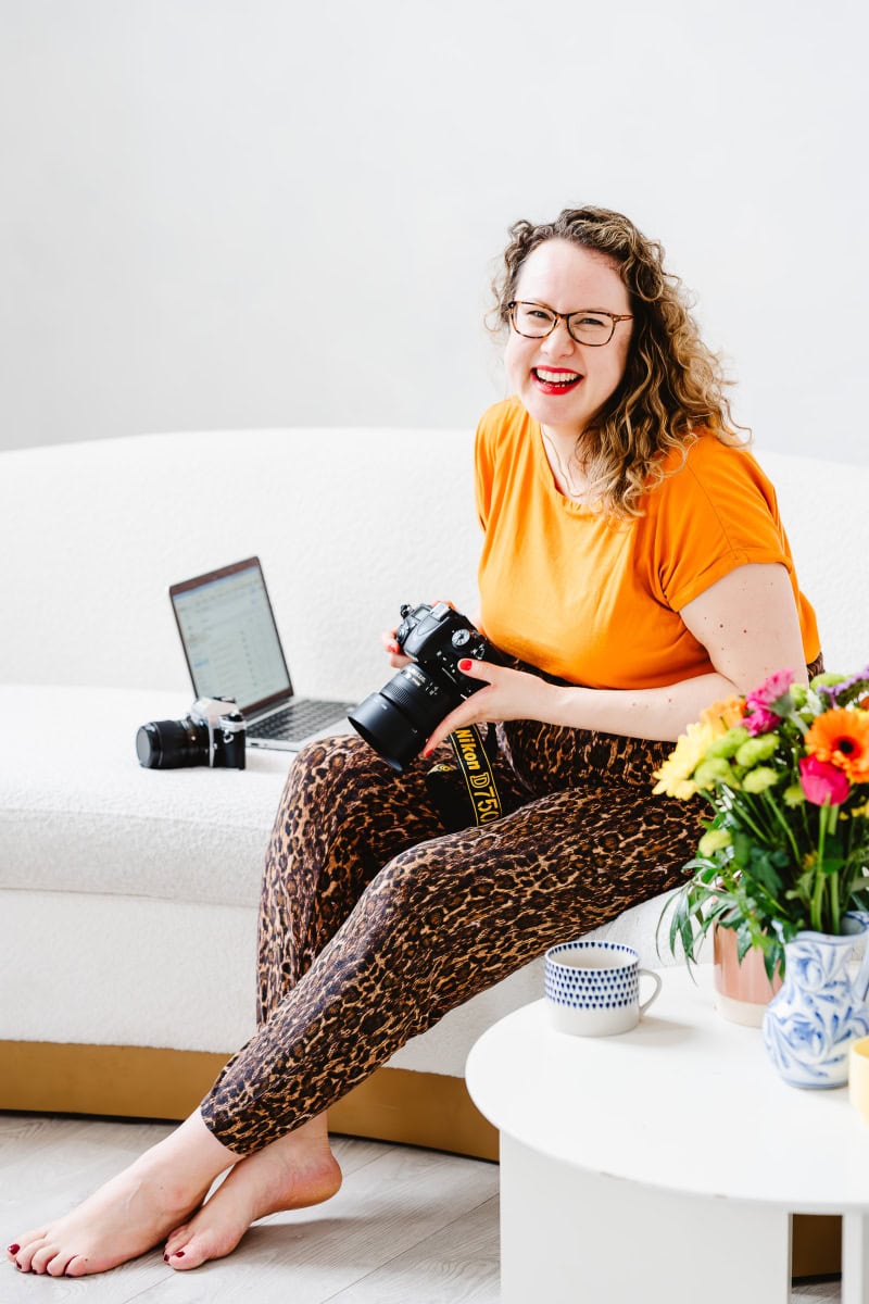 A portrait photograph of Molly Rhianon Behagg the founder of Molly Rhianon Photography. Molly sits on a white sofa holding a camera and laughing. She has curly hair and wears an orange top and leopard print trousers. 