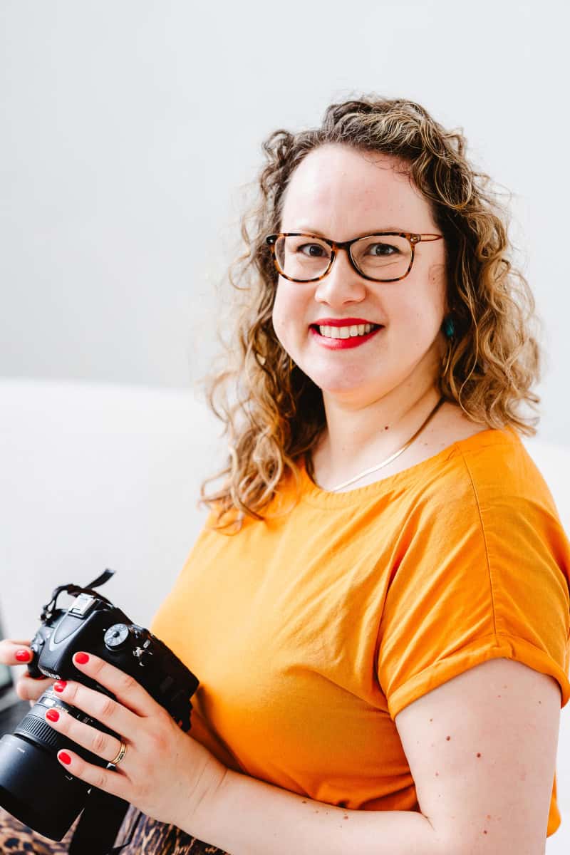 Molly rhianon looking at the camera smiling. she holds a camera in her hand and wears an orange top