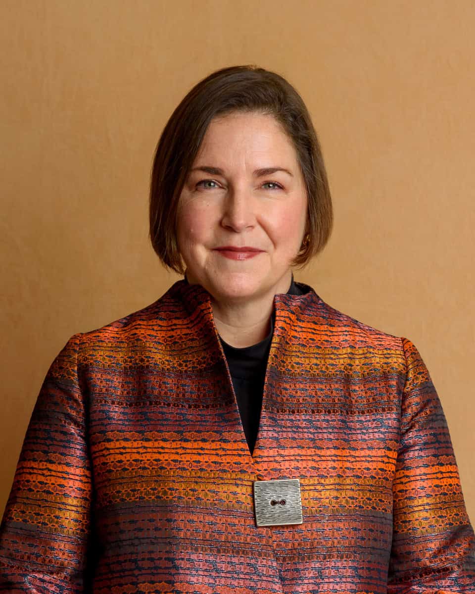 A professional headshot of Dame Nicole Jacobs the Domestic Abuse Commissioner taken by Molly Rhianon Photography. Nicole has short brown hair and is smiling against a tan backdrop and wearing an orange and purple patterned jacket.